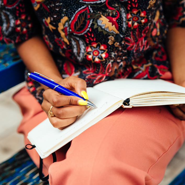 a girl sitting having a notepad on her lap and holding a pen 